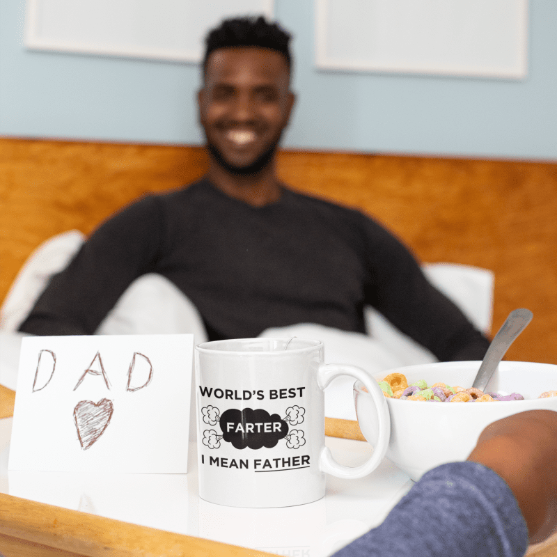 Vader en kind lachen samen, terwijl een koffiemok met de tekst 'World's Best Farter, I Mean Father' op tafel staat - Leuk Vaderdagcadeau"